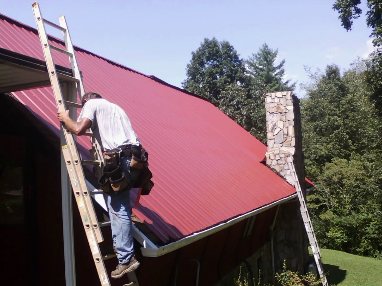 Roofer on a ladder beside a red metal roof and stone chimney — Soffit & Fascia Repair by Roofing Brothers in Browder